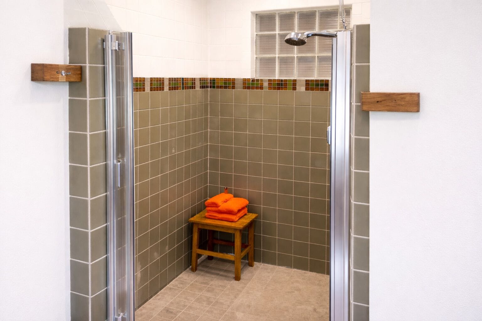 A shower with a glass door, a wooden stool with orange towels, and a tiled wall with a colorful border.