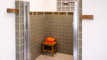 A shower with a glass door, a wooden stool with orange towels, and a tiled wall with a colorful border.