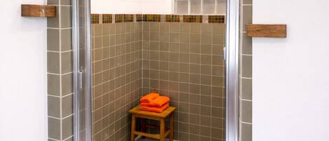 A shower with a glass door, a wooden stool with orange towels, and a tiled wall with a colorful border.