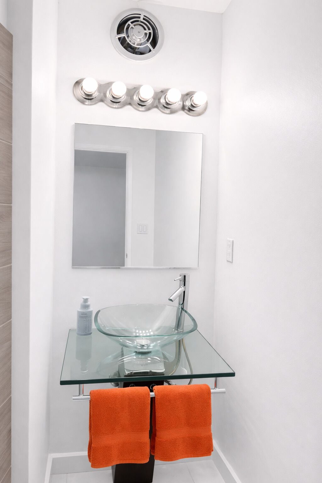 Une salle de bain avec un lavabo en verre, deux serviettes orange, un miroir et une bouche d’aération au plafond.
