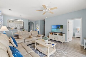 Ein Wohnzimmer mit Sofa, Couchtisch und Fernseher, im Hintergrund eine Küche und ein Deckenventilator.