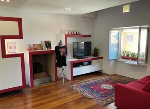 A modern living room with a flat-screen TV, a red sofa, a patterned rug, and a window with a view of greenery.