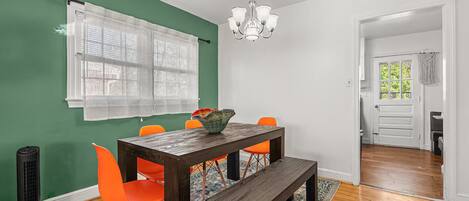 Un coin repas avec une table en bois, des chaises orange, un banc, un lustre et un tapis à motifs.