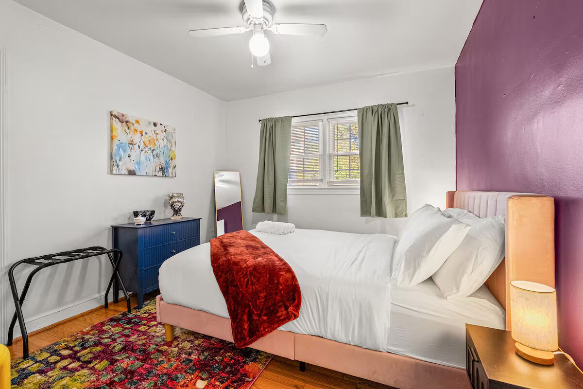 A bedroom with a bed, a desk, a chair, a ceiling fan, a window with curtains, and a colorful rug.