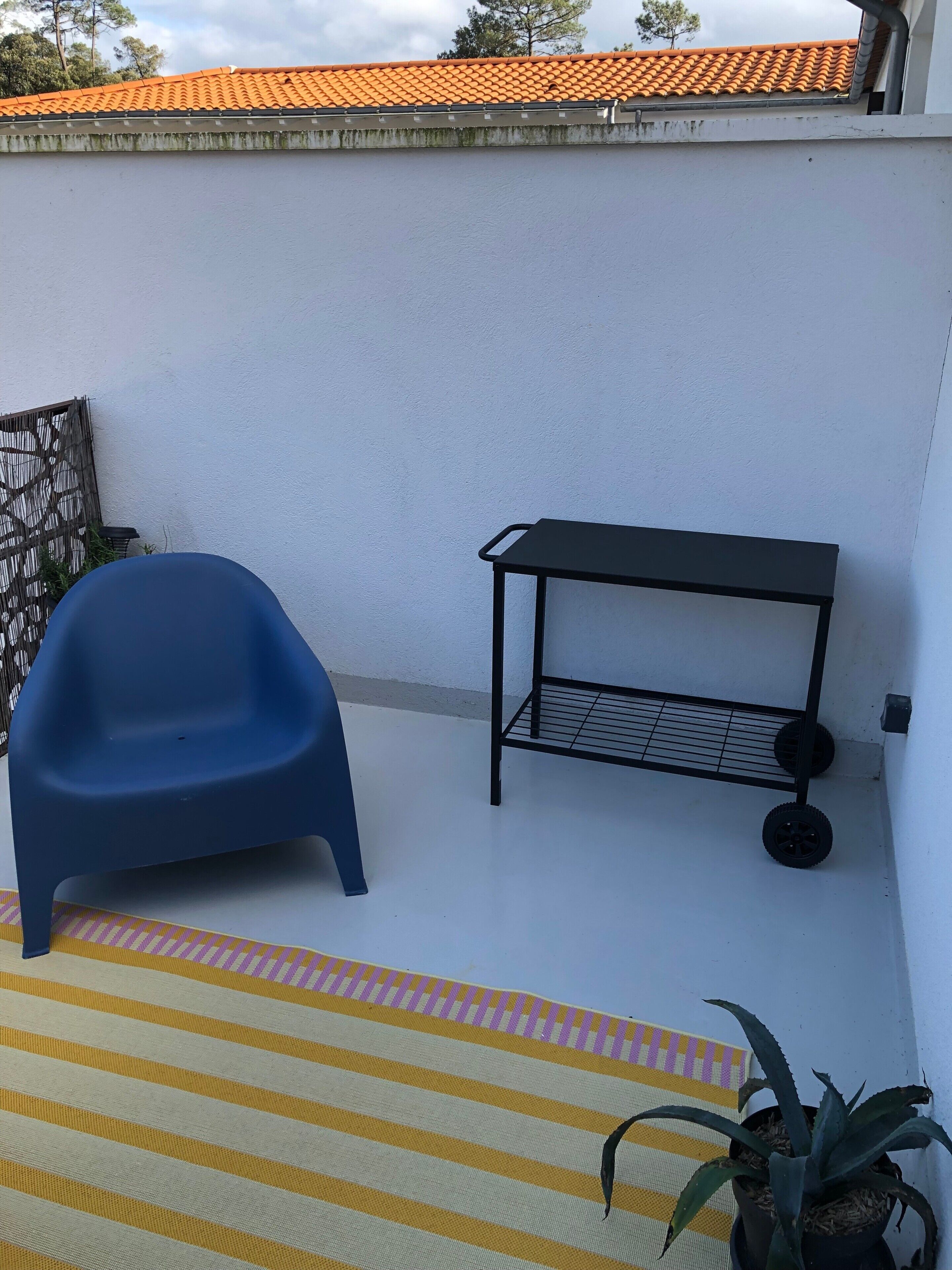 Une chaise en plastique bleue, une étagère en métal et un tapis à rayures sur une terrasse sur le toit.