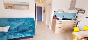 Un espace de vie compact avec un canapé bleu, une petite kitchenette et une vue sur la plage depuis la porte.