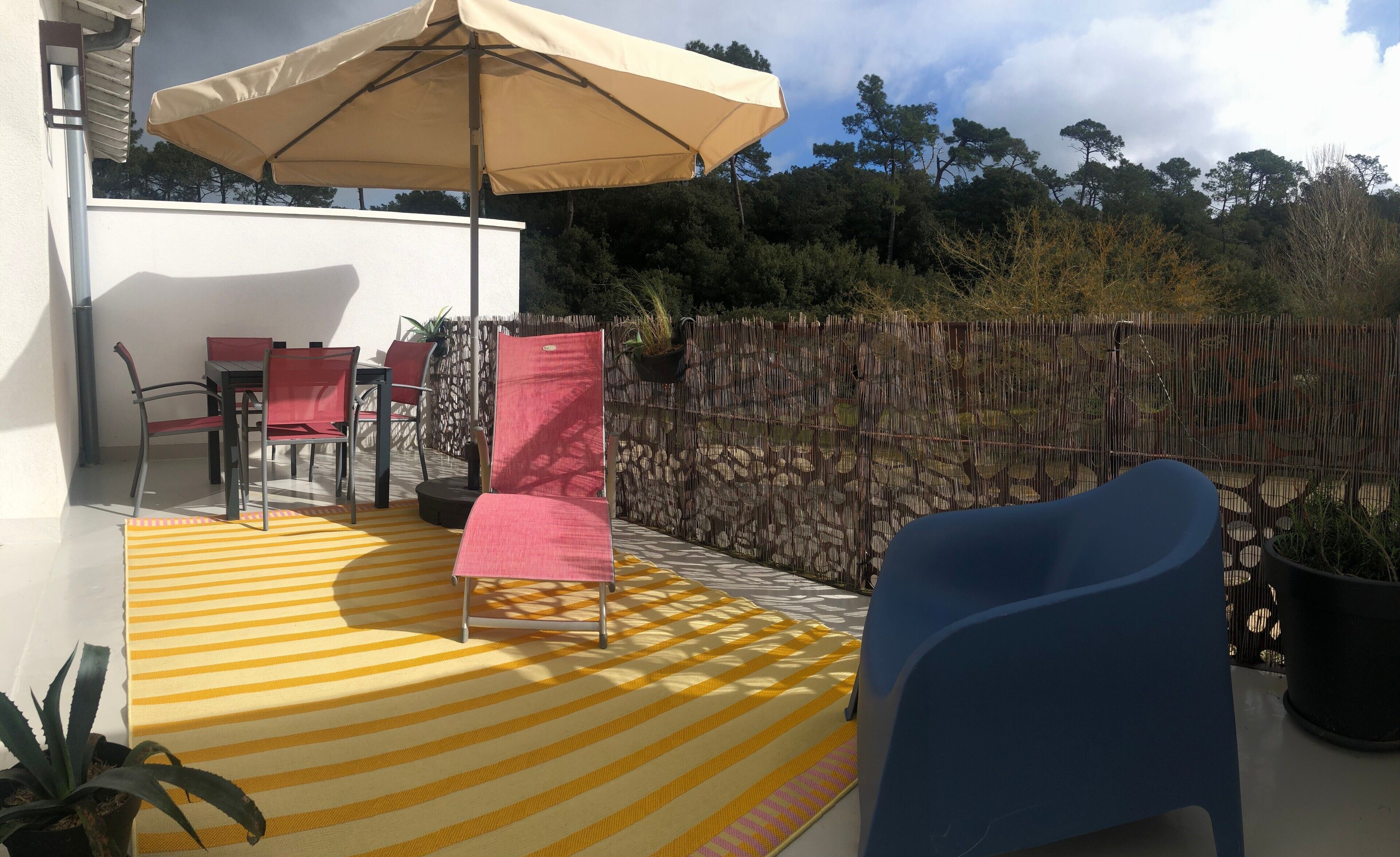 Eine Terrasse mit einem gelb-gestreiften Teppich, einem blauen Stuhl, einem roten Stuhl und einem beigefarbenen Sonnenschirm.