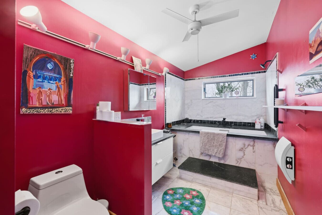 A bathroom with a red wall, a white toilet, a sink, a mirror, and a ceiling fan.