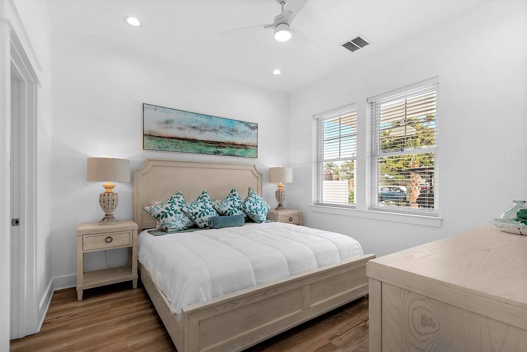 Une chambre à coucher comprenant un lit, deux tables de chevet, un ventilateur de plafond, une fenêtre avec des stores et un tableau accroché au mur.