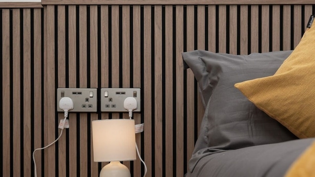 Une chambre à coucher avec une tête de lit en bois, une table de chevet avec une lampe et un lit recouvert d’une couverture grise et agrémenté d’un oreiller jaune.
