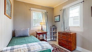 A bedroom with a bed, a dresser, a lamp, a shelf, and a window with curtains.