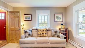 A living room with a beige sofa, two windows, a red door, and a small wooden side table with a lamp.