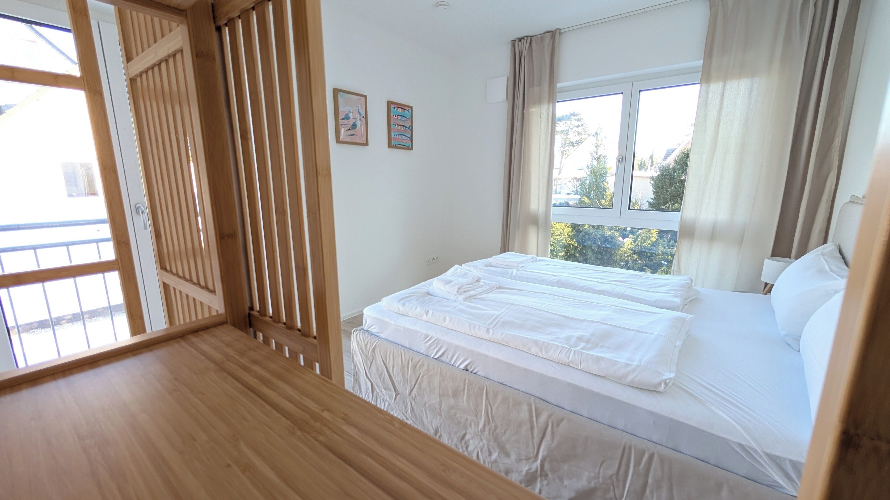 Ein Schlafzimmer mit Holzboden, einem Bett mit weißen Leinentüchern, einem Fenster mit Vorhängen und einer Wand mit zwei gerahmten Bildern.