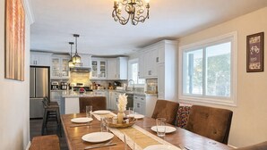 A dining area with a wooden table set for four, white cabinets, a chandelier, and a window with a view. - River Retreat- Hot tub/Games/BBQ/Bonfire/Sleeps 14 (Tweed)