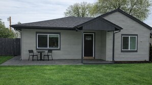 A single-story house with a grey exterior, a front door, and a small patio with two chairs and a table. - Welcome to the Sundancer @ the Little Littleton Village (Littleton)