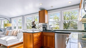 A modern kitchen with wooden cabinets, a stainless steel dishwasher, and a large window with a view of trees. - Harbor Views, Parking, Roof Deck, Beach - West End (Provincetown)