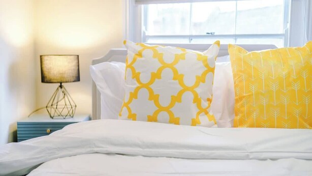 A bed with white and yellow pillows, a yellow blanket, a bedside lamp, and a window with a view. - Charming Grade II Listed Apartment | Sleeps 4 (Wem)