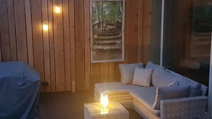 A cozy outdoor seating area with a wicker sofa and a covered table, a framed picture of a staircase, and a lantern on a square table.