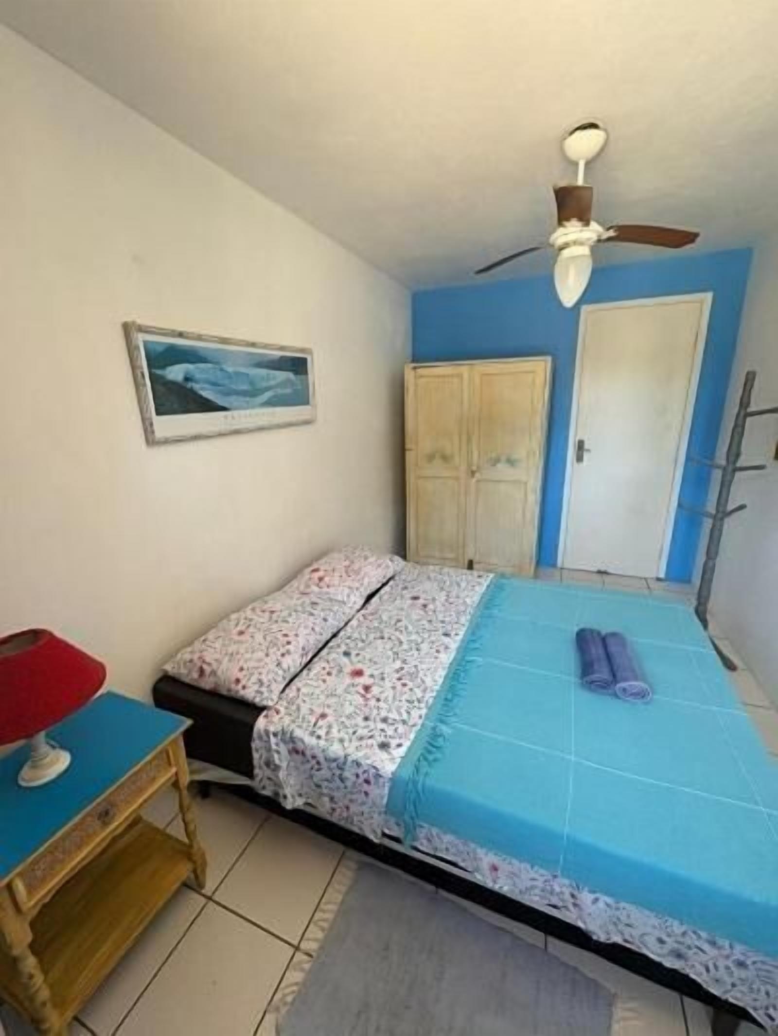 A bedroom with a bed, a ceiling fan, a wooden wardrobe, a door, and a small table with a red chair.