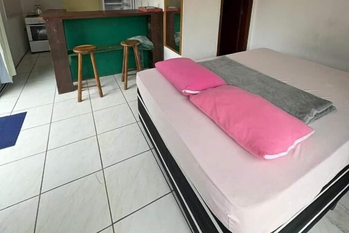 A compact room with a bed, a small stool, a desk, and a kitchenette area.
