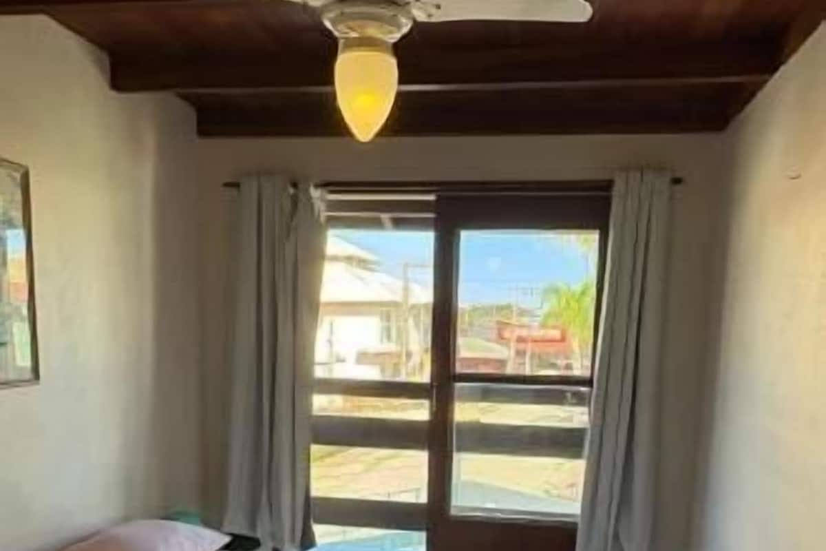 A single bed with a white and pink bedspread in a room with a ceiling fan, a window with curtains, and a view of buildings outside.
