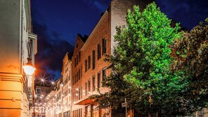 Una calle iluminada de noche con edificios, un árbol y farolas.