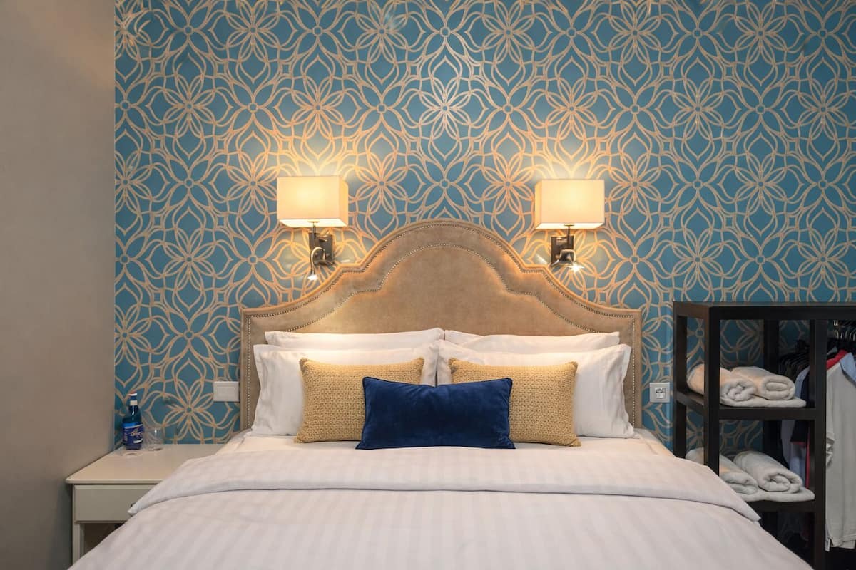 A hotel room with a patterned wallpaper, a bed with white linens and blue and beige pillows, two wall-mounted lamps, and a nightstand with a bottle and glasses.