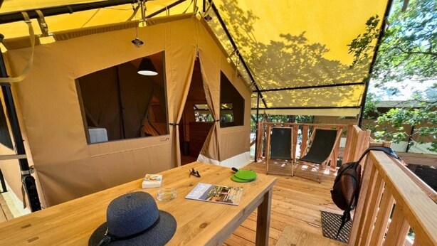 A tented accommodation with a wooden deck, a table with a hat and a magazine, and a view of the outdoors. - ZEcamping 3 étoiles animé piscine chauffée locatif 20m2 5 pers (Saint-Saturnin-lès-Apt)