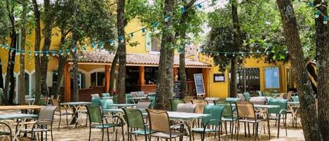 An outdoor cafe with tables and chairs, surrounded by trees and a yellow building.