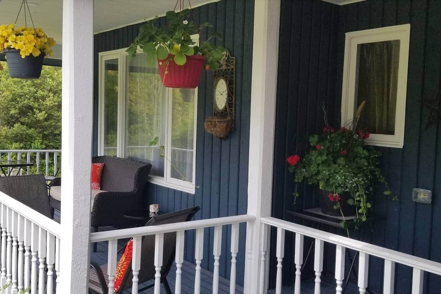 A porch with a blue exterior, white railing, and two chairs. There are hanging plants and a wall clock.