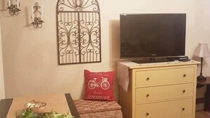 A living room with a wooden table, a television on a stand, a decorative wall piece, and a small sofa with a cushion. - The Garden Room, Trinity Guest House (Trinity)