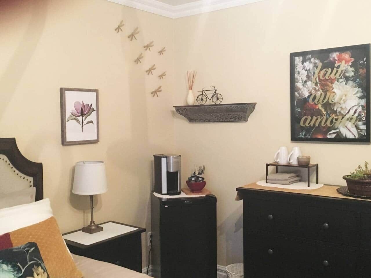 Una habitación de hotel con una cama, una mesita de noche con lámpara, una cafetera, un estante con adornos y un cuadro en la pared.