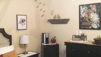 Une chambre d’hôtel comprenant un lit, une table de chevet avec une lampe, une machine à café, une étagère avec des objets décoratifs et un tableau encadré au mur.
