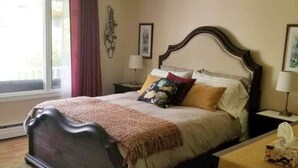 A bedroom with a large bed, a bedside table, a lamp, and a window with curtains. - The Garden Room, Trinity Guest House (Trinity)