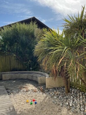 A backyard with a wooden deck, a concrete planter with a palm tree, and a gravel area with colorful balls. - Dunes Walk Cottage -  a cottage that sleeps 4 guests  in 2 bedrooms (Camber)
