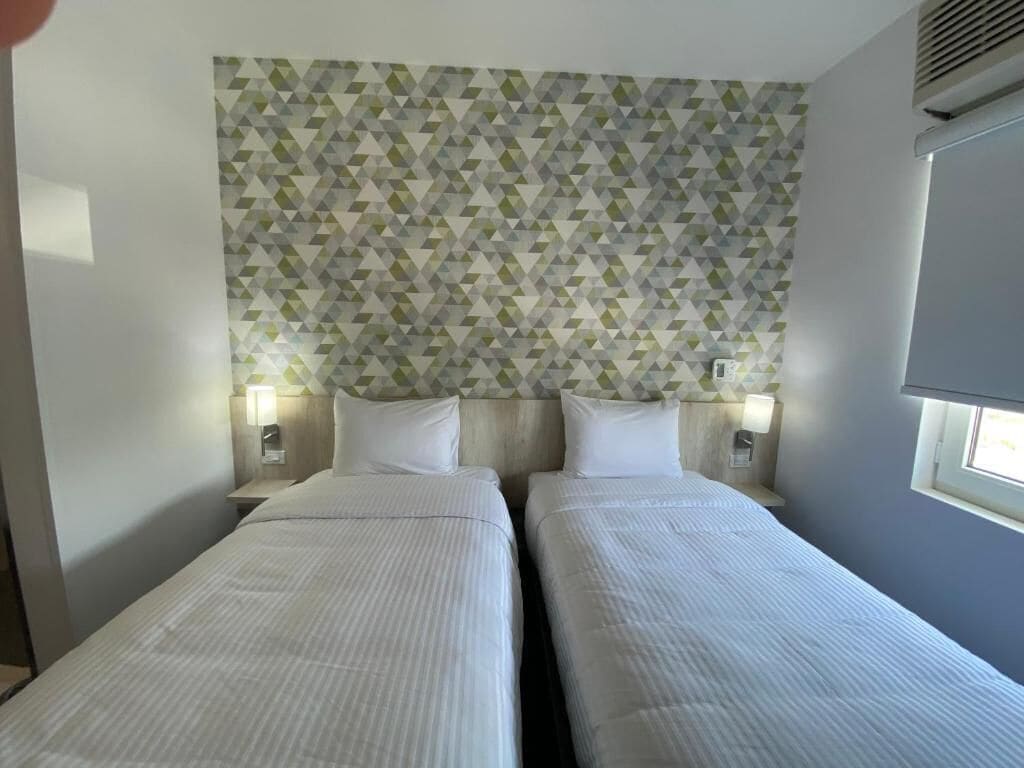 Habitación de hotel con dos camas, un diseño de pared geométrico, lámparas de noche y una ventana con persianas.