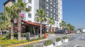 A modern hotel with a prominent 'hi!' sign, a covered outdoor seating area, and a parking lot with cars. - HI MONTERREY AEROPUERTO (Apodaca)