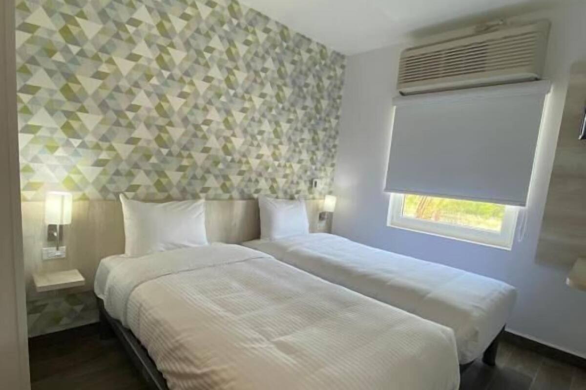 Camera d'albergo con due letti, parete decorata con un motivo geometrico, finestra con persiane e condizionatore d'aria a parete.