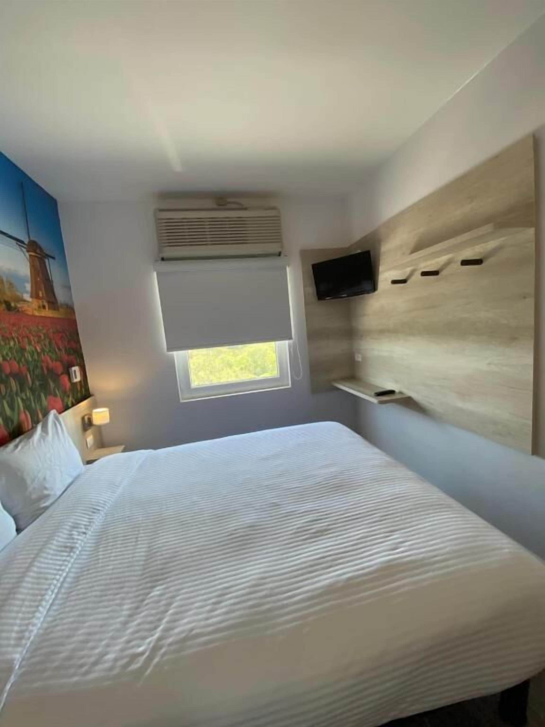 A modern hotel room with a large bed, a mounted TV, a window with blinds, and a painting on the wall.