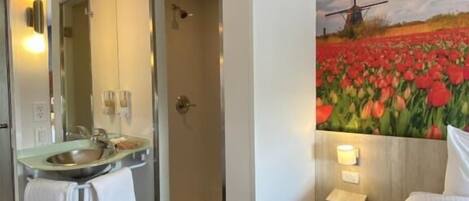 Habitación de hotel con baño que incluye un lavamanos, espejo y ducha. Hay una cama con cabecero y una lámpara de pared.