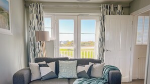 Ein gemütliches Wohnzimmer mit einem dunkelgrauen Sofa, mehreren Kissen und einer türkisblauen Decke. Es gibt zwei Fenster mit Vorhängen, eine Tür und eine Stehlampe.