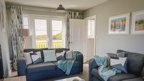 Ein Wohnzimmer mit zwei grauen Sofas, einem Couchtisch mit Buch, Teekanne und Tassen, und Blick auf einen Balkon durch große Fenster.