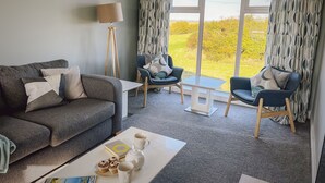 Ein modernes Wohnzimmer mit einem grauen Sofa, zwei Sesseln, einem Couchtisch und einer Stehlampe. Große Fenster mit gemusterten Vorhängen bieten Blick auf Grünflächen und ein entferntes Gebäude.