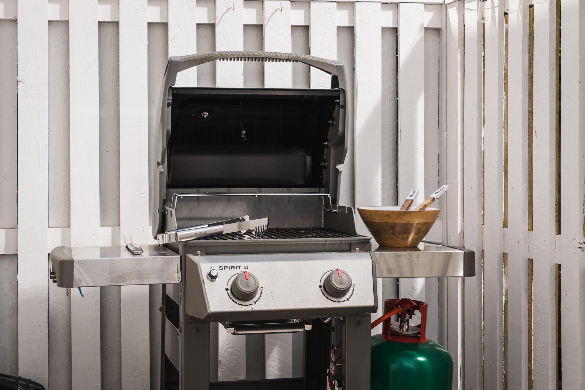 A portable gas grill with two control knobs, a metal grate, and a wooden bowl with utensils on a metal stand.
