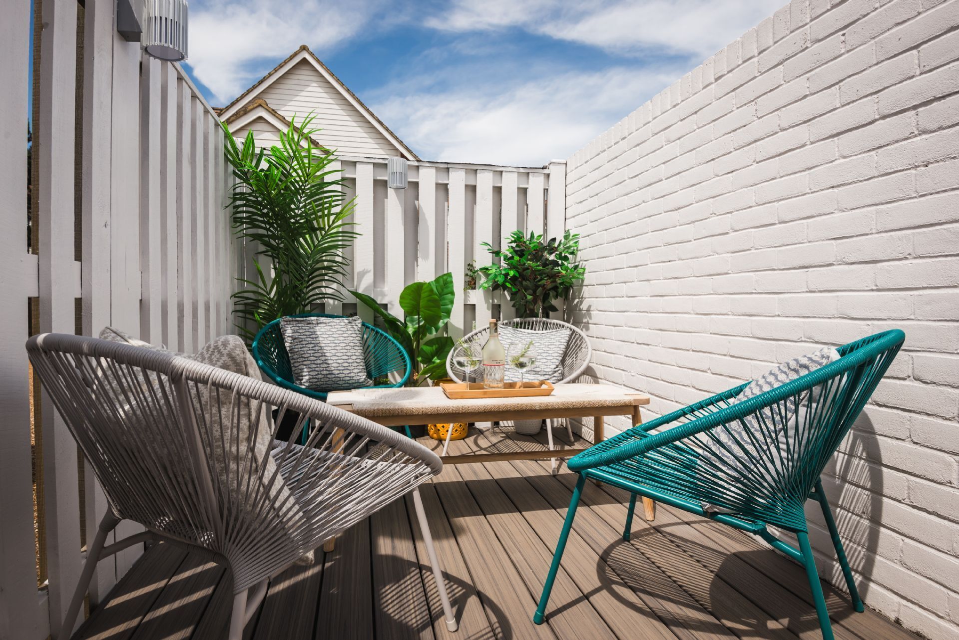 A wooden deck with wicker chairs and a table, surrounded by white fences and potted plants.
