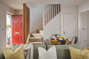 A living room with a sofa set, a dining table with breakfast items, a red door, and a staircase. - Tradewinds -  a cottage that sleeps 4 guests  in 2 bedrooms (Camber)