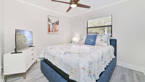 Une chambre à coucher comprenant un lit, un ventilateur de plafond, une fenêtre avec des stores, une table de chevet avec une lampe et un téléviseur posé sur un meuble.