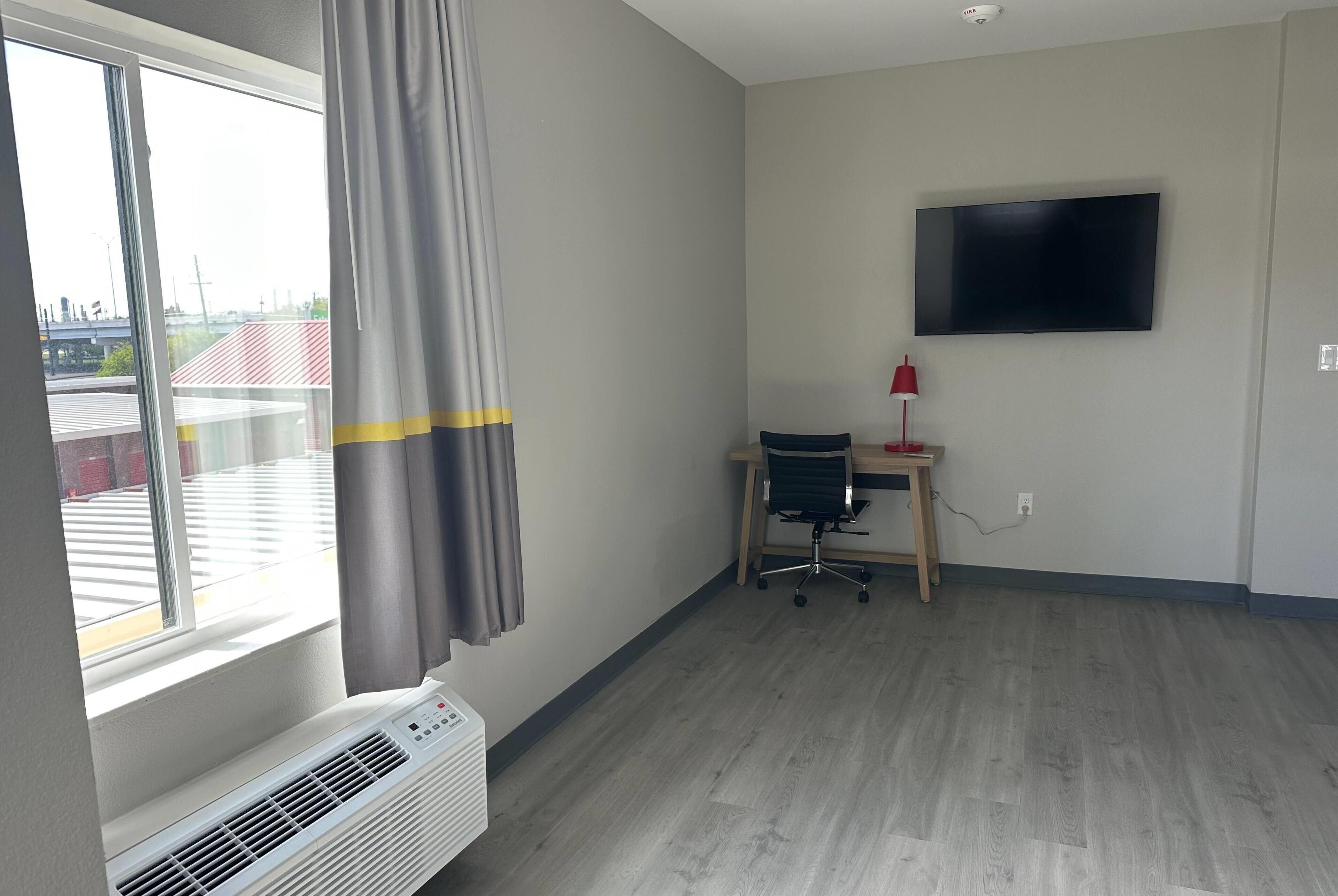 Quarto com uma janela grande, uma escrivaninha com cadeira, televisão de tela plana e abajur vermelho.