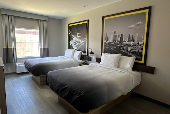 A hotel room with two beds, a window with curtains, a cityscape painting, and a wall-mounted lamp.