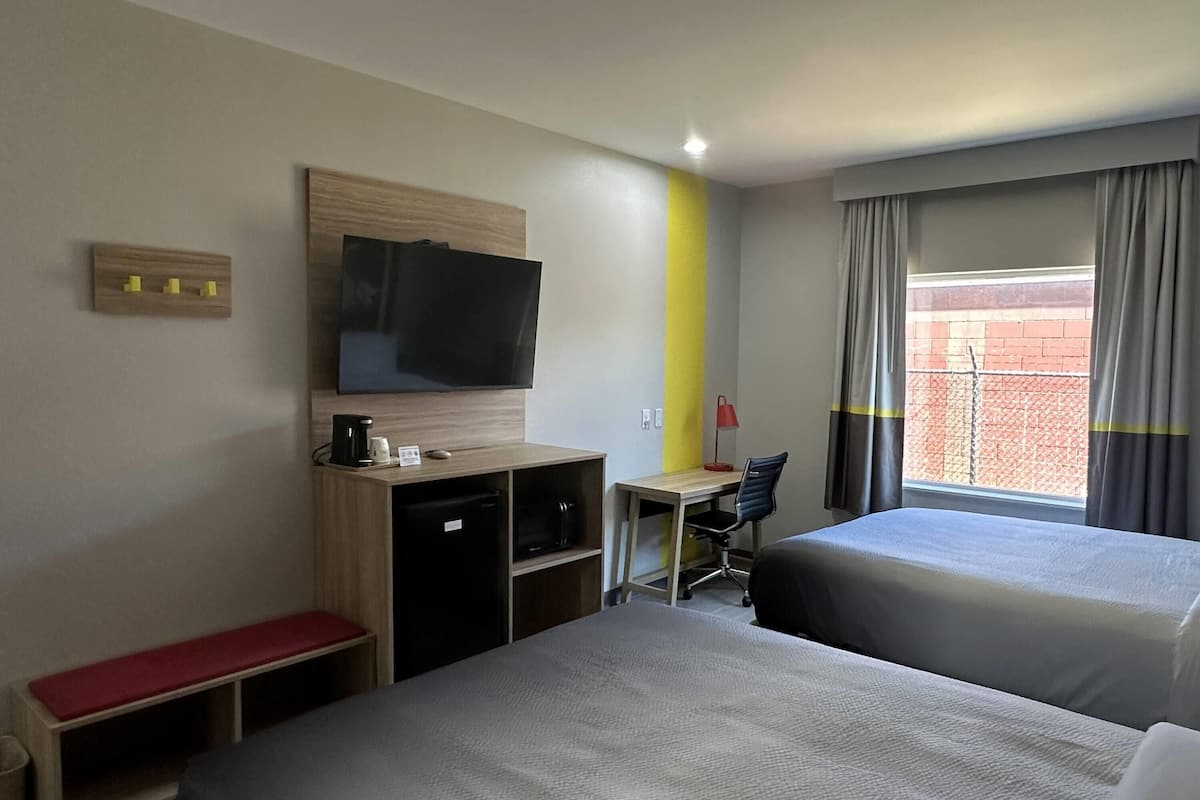 Una habitación de hotel con dos camas, un escritorio, un televisor, una cafetera y un ventanal con cortinas.